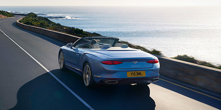 Bentley Guatemala New Bentley Continental GTC Azure convertible rear view in Light Windsor blue paint with 22 inch ten spoke wheels driving dynamically on an ocean road in the sunshine